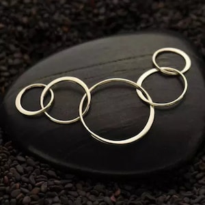 May include: A silver necklace with five interlocking circles of varying sizes. The necklace is displayed on a black stone surface.