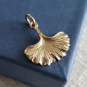 May include: A polished gold-tone metal pendant shaped like a ginkgo leaf. The charm features a fan-like silhouette with delicate, radiating ridges and a smooth, curved stem that connects to a small circular jump ring at the top for attachment.