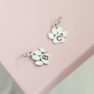 May include: Two silver-toned metal charms shaped like animal paw prints. Each charm features a small jump ring at the top for attachment. One paw print charm is engraved with the uppercase letter B, and the other is engraved with the letter C.