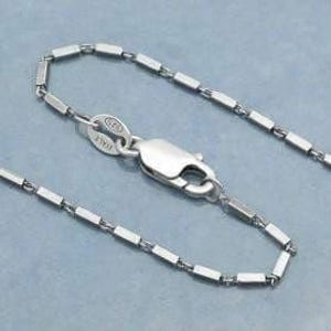 May include: A close-up of a silver necklace with a rectangular link chain and a lobster clasp. The clasp is connected to a small tag with the text "925 ITALY". The necklace is displayed on a light blue surface.