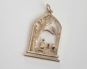 May include: A gold-toned metal pendant shaped like an arched window frame. The design features a bare tree with a bird perched on a branch, overlooking three gravestones set within the frame. A small circular bail is attached to the top.