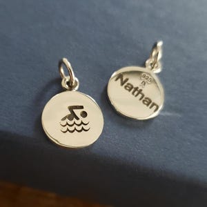 May include: Two circular silver charms with jump rings. One charm features a cutout of a person swimming in waves. The other charm is engraved with the name Nathan and the hallmark 925, indicating sterling silver composition and quality.