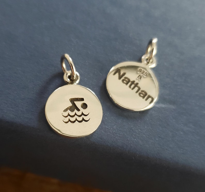 May include: Two circular silver charms with jump rings. One charm features a cutout of a person swimming in waves. The other charm is engraved with the name Nathan and the hallmark 925, indicating sterling silver composition and quality.