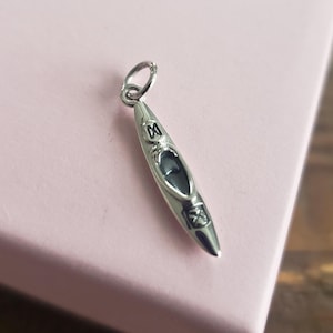 May include: A silver charm shaped like a kayak. The charm features a detailed design and a loop at the top for attaching to a bracelet or necklace.