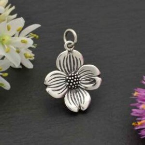 May include: A silver charm in the shape of a four-petaled flower with a textured surface. The center of the flower has a small, raised circle with a textured surface.