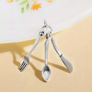 Peut inclure: Un charm en métal argenté composé de trois ustensiles miniatures : une fourchette, une cuillère et un couteau dentelé. Les pièces sont reliées en haut par un petit anneau circulaire, formant un pendentif suspendu à la finition métallique polie.