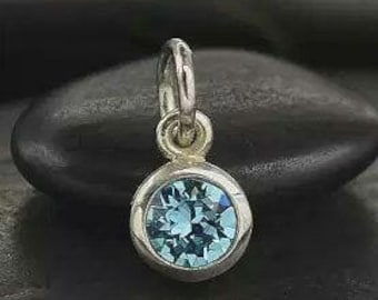May include: A silver pendant featuring a round, light blue gemstone. The gemstone is set in a bezel setting and has a faceted appearance. The pendant is suspended from a small loop.