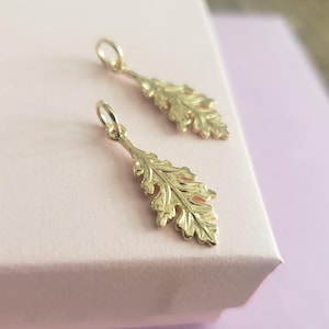 May include: Two gold-toned metal charms shaped like detailed oak leaves. Each charm features a textured surface with defined veins and a small circular jump ring at the top for attachment. The charms have a polished finish and reflective surface.