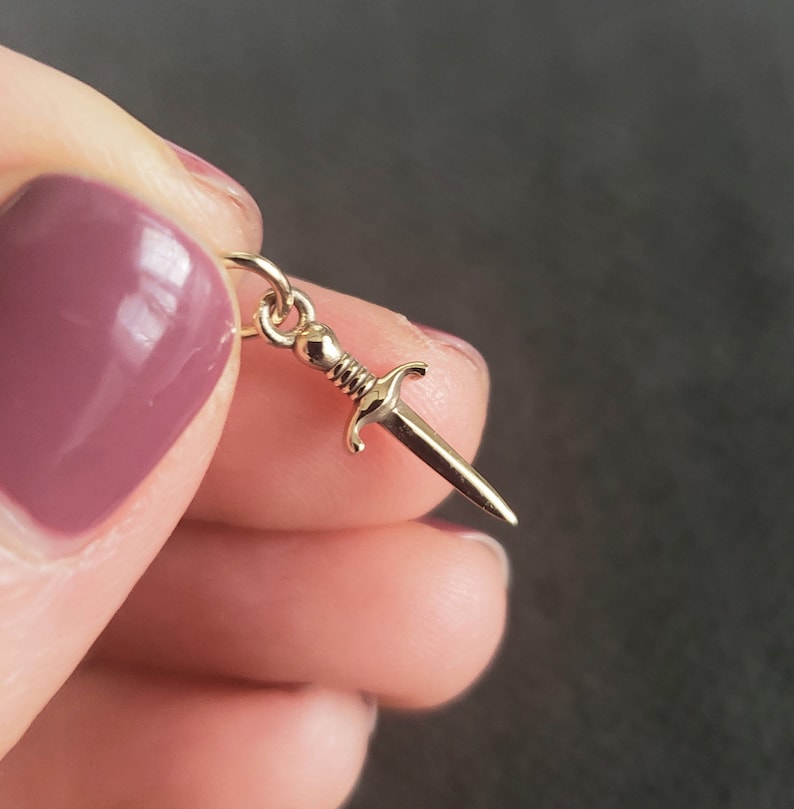 May include: A gold-colored dagger charm pendant. The charm is detailed with a pointed tip and a cross guard.