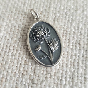 May include: Oval silver pendant featuring a detailed floral design. The pendant showcases a dark gray background with a raised relief of a chrysanthemum and smaller flower, along with leaves. The pendant has a silver bail for a chain.
