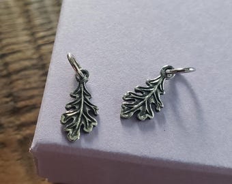 May include: Two small, silver-colored oak leaf charms. Each charm has detailed leaf veins and a small loop at the top for attaching to a chain or bracelet. The charms are displayed on a light purple surface.