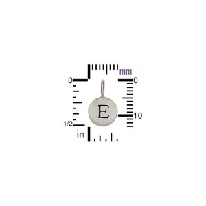 以下が含まれることがあります： 丸い銀色のペンダントで、表面に文字「E」が刻まれています。チェーンを取り付けるための小さなループが付いています。ペンダントの直径は約1 cmです。画像には、インチとミリメートルで測定値を示す定規が含まれています。