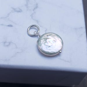 May include: A round, iridescent pearl pendant with a silver loop. The pearl has a flat, disc-like shape and a subtle, pearlescent sheen. The pendant is likely designed for use as a necklace charm or earring component.
