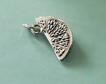 May include: A silver-colored charm in the shape of a citrus fruit slice. The charm has detailed texturing on the inside, with a small loop for attaching to a necklace or bracelet. The charm is set against a light blue background.