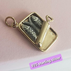 Può includere: Un ciondolo in metallo dorato a forma di scatoletta rettangolare aperta contenente due pesci argentati testurizzati. Il ciondolo presenta un piccolo anellino e una linguetta triangolare laterale. Il testo 925 sterling silver è visibile.