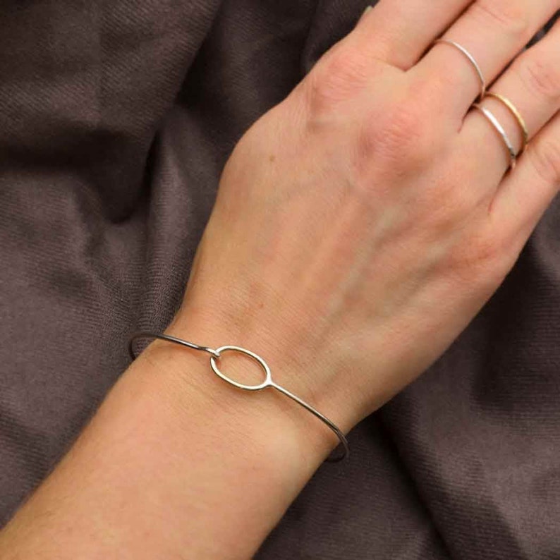 以下が含まれることがあります： シンプルな楕円形のデザインのシルバーバングルブレスレット。ブレスレットは細くて繊細で、人の手首につけられています。