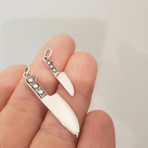 May include: Two silver knife charms, one larger than the other, with detailed handles. The charms are held in a hand.