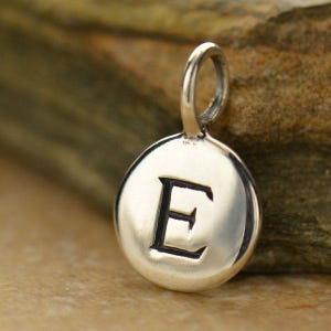 以下が含まれることがあります： 太字のセリフ体で「E」の文字が刻まれたシルバーペンダント。円形のペンダントは研磨仕上げで、チェーン用の小さなループが付いています。背景はぼやけた天然石です。