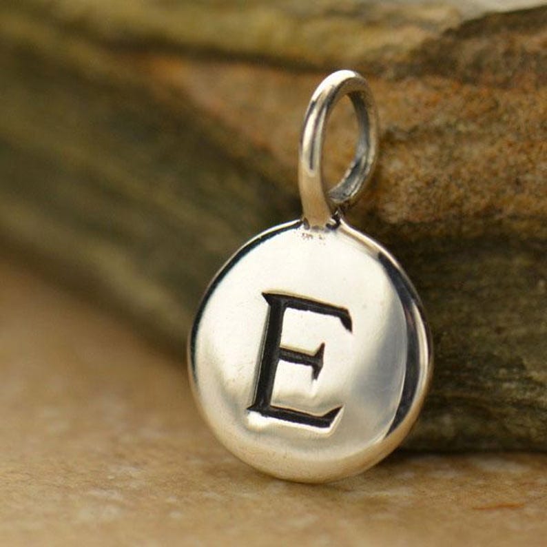 以下が含まれることがあります： 太字のセリフ体で「E」の文字が刻まれたシルバーペンダント。円形のペンダントは研磨仕上げで、チェーン用の小さなループが付いています。背景はぼやけた天然石です。