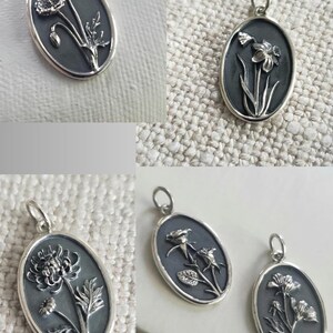 May include: Oval-shaped silver pendants with floral designs. Each pendant features a different flower, including a poppy, daffodil, rose, and other blooms. The pendants have a dark background and a silver border, with a loop at the top for a chain.