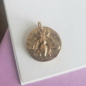 May include: A gold-colored bee pendant with a detailed bee design. The pendant is circular with a textured background and a small loop at the top for a chain. The bee has detailed wings and body.