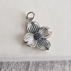May include: A silver charm in the shape of a dogwood flower. The flower has five petals and a center with a textured design.