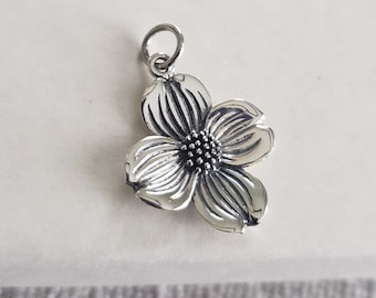 May include: A silver-toned metal charm shaped like a four-petaled dogwood flower. The petals feature embossed linear detailing radiating from a central cluster of small, raised dots. A circular jump ring is attached to the top of the flower.