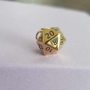 Könnte beinhalten: Ein goldfarbener Metallanhänger in Form eines zwanzigseitigen Würfels, auch d20 genannt, mit eingravierten schwarzen Zahlen wie 20, 8, 10, 12, 14 und 16. Der geometrische polyedrische Anhänger hat einen kleinen runden Ring an der oberen Ecke.