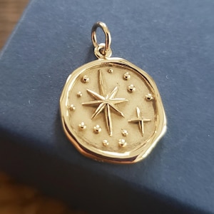 May include: Gold pendant featuring a star with a smaller star and dots around it, set in a round, textured frame.