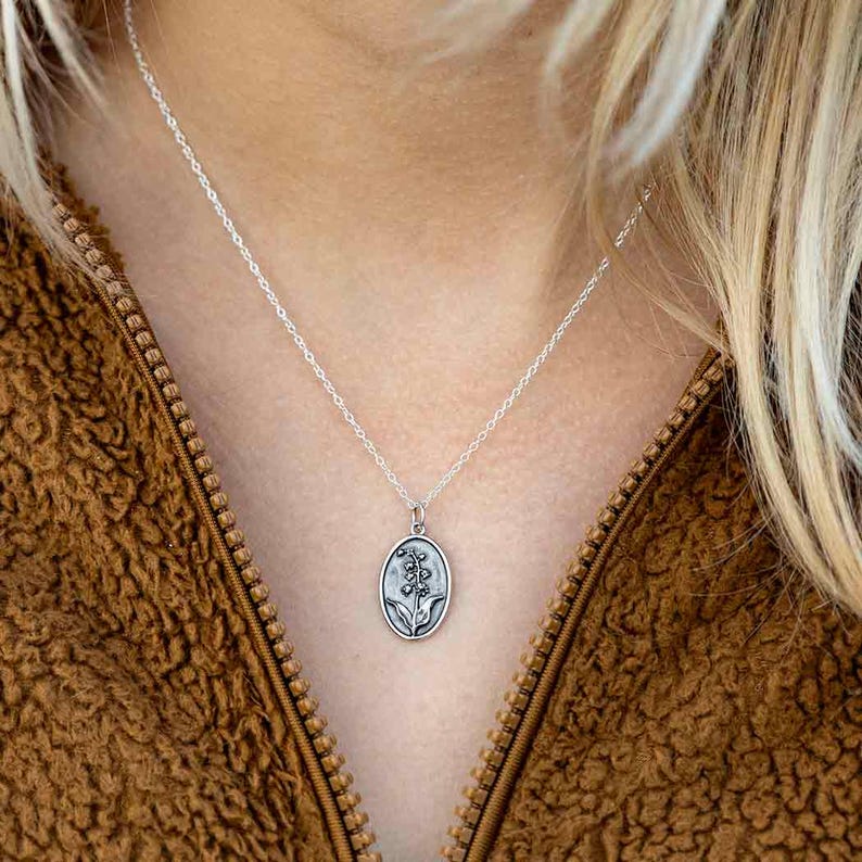 May include: Silver-toned necklace with an oval pendant featuring a floral design. The pendant hangs from a delicate chain. The necklace is worn against a brown, textured jacket.
