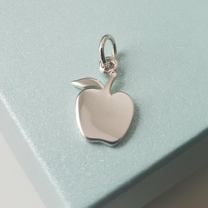 Puede incluir: Un amuleto de plata con forma de manzana con una hoja. El amuleto tiene un bucle en la parte superior para sujetarlo a un collar o pulsera.