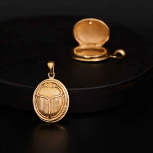 May include: A gold-toned oval pendant featuring a raised scarab beetle design on the front, accompanied by a matching open locket pendant. Both pieces have a polished metallic finish and include a small loop at the top for attachment to a chain.