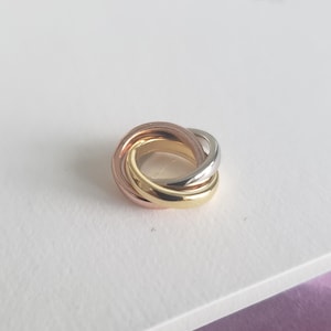 Könnte beinhalten: Ein Ring mit drei ineinandergreifenden Bändern in Roségold, Gelbgold und Silber. Die Bänder sind glatt und poliert und bilden ein nahtloses, ineinander verschlungenes Design. Der Ring ist ein klassisches Schmuckstück.