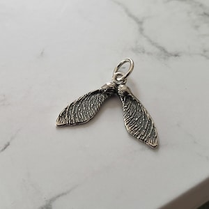 Puede incluir: Encanto de plata en forma de dos semillas de arce. El encanto está detallado con una superficie texturizada.