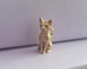 May include: A gold-colored cat charm in a sitting position. The charm has detailed features, including pointed ears, a textured coat, and a small loop for attachment. The cat is set against a soft, light purple background.