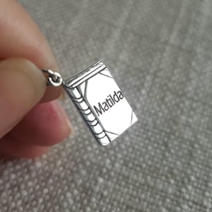 May include: A small, silver-colored pendant in the shape of a book. The word "Matilda" is engraved on the cover. The pendant is detailed with lines to represent the pages and spine of the book. The pendant is attached to a small loop.