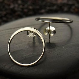 May include: A pair of silver-colored, open-circle stud earrings. The earrings are circular with a post and butterfly backing. The earrings are displayed on a dark, textured surface. The earrings are a simple, modern design.
