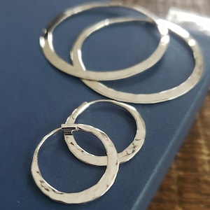 May include: A set of silver-toned hoop earrings with a hammered texture. The earrings are displayed on a dark blue surface. Two larger hoops are positioned behind a pair of smaller hoops, creating a layered effect.