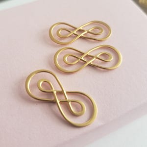 May include: Three gold-coloured infinity symbol charms on a pale pink surface. The charms are made of thin metal wire with a polished finish. The infinity symbols are a popular design element for jewellery and crafts.