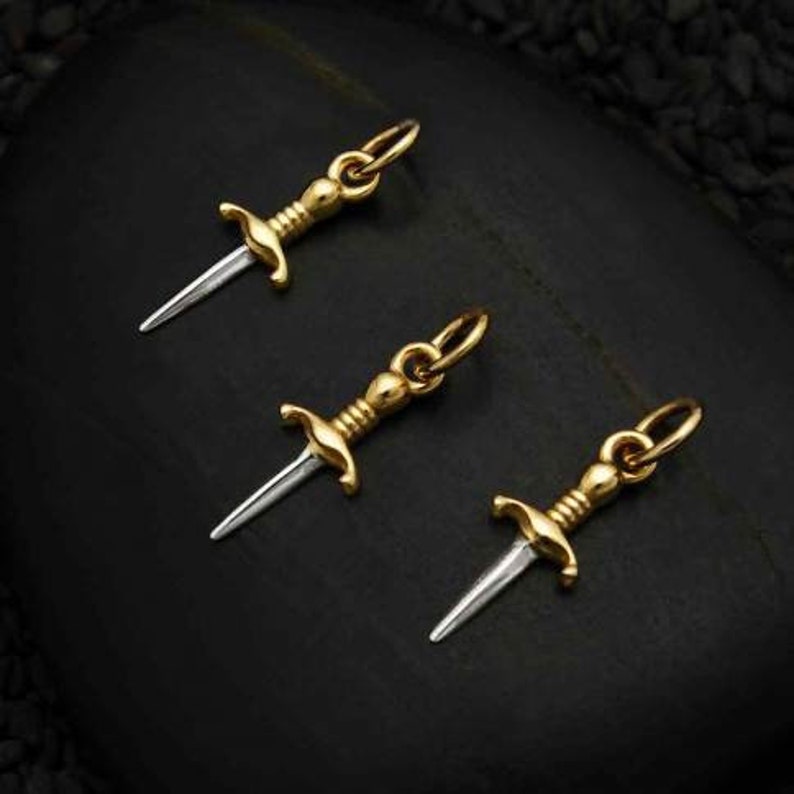 May include: Three gold-toned dagger charms with silver blades. The charms are on a black background.