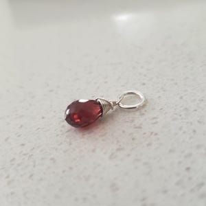 May include: A teardrop-shaped, deep red garnet pendant with a silver wire wrap and a silver loop. The gemstone is a rich, translucent colour. The pendant is designed for use as a necklace charm.