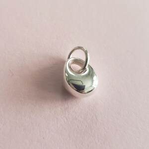 May include: A small, polished silver pendant with a smooth, rounded form. It features a loop at the top for a chain. The pendant's reflective surface catches the light, creating highlights and shadows. The background is a soft pink.