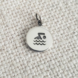 May include: A circular silver charm featuring a black engraved icon of a person swimming in water. The charm has a small jump ring attached to the top, allowing it to be suspended from a chain or bracelet. The surface of the metal is polished and smooth.