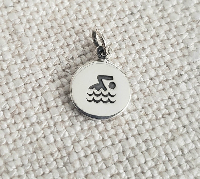 May include: A circular silver charm featuring a black engraved icon of a person swimming in water. The charm has a small jump ring attached to the top, allowing it to be suspended from a chain or bracelet. The surface of the metal is polished and smooth.