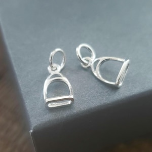 May include: Two silver stirrup charms, each with a loop for attaching to a necklace or bracelet.