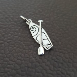 K&ouml;nnte beinhalten: Ein silberfarbener Anh&auml;nger mit einem Stand-Up-Paddleboard und Paddel. Das Paddleboard hat ein Spiraldesign und eine sternf&ouml;rmige Form. Der Anh&auml;nger hat eine kleine Schlaufe zum Befestigen an einer Kette oder einem Armband.