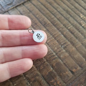 May include: A small, circular silver-toned metal charm featuring a black engraved icon of a person swimming in water. The charm includes a small jump ring attached to the top, allowing it to be easily connected to a necklace chain or bracelet.