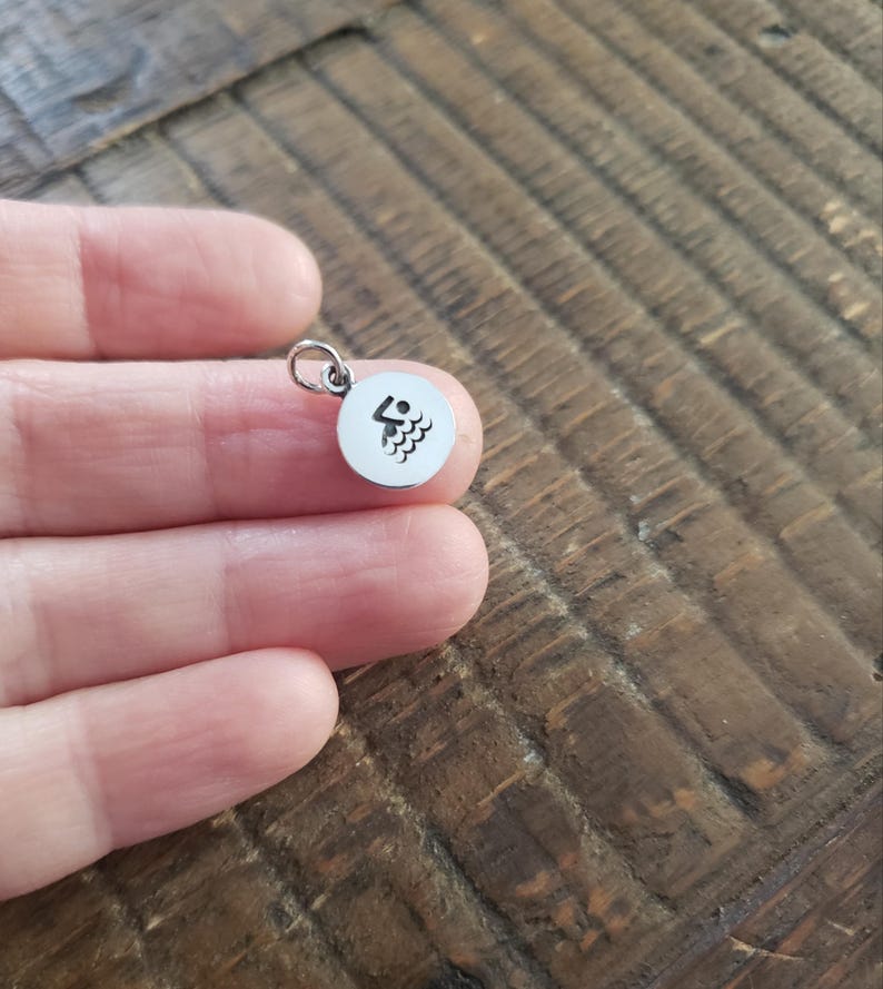 May include: A small, circular silver-toned metal charm featuring a black engraved icon of a person swimming in water. The charm includes a small jump ring attached to the top, allowing it to be easily connected to a necklace chain or bracelet.