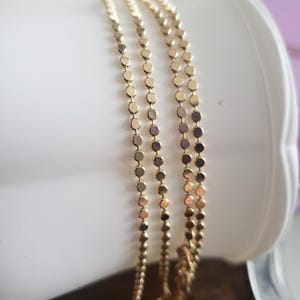 May include: Gold-colored necklace with a delicate design. The necklace features multiple strands of small, flat, circular links. The jewelry is draped over a white surface, showcasing its texture and shine. The necklace is a simple yet elegant accessory.