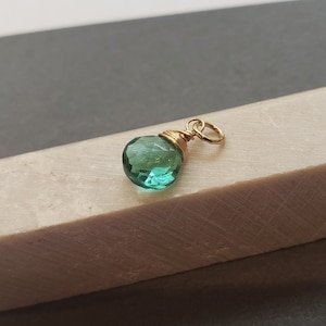 May include: A faceted, teardrop-shaped green gemstone pendant secured with gold-colored wire wrapping. The pendant features a small, circular gold-colored jump ring attached to the top, allowing it to be suspended from a chain or cord.